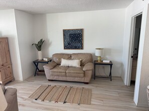 Un salon avec un canapé beige, un tapis à motifs, un élément décoratif mural et une petite table avec une lampe.