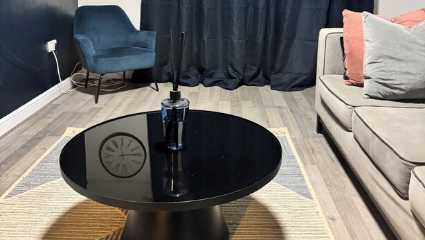 A modern living room with a round black coffee table, a blue armchair, a beige sofa with pillows, a dark curtain, and a wall clock. - Corporate 3bedroom home with parking (Erith)