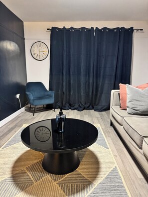 Una sala moderna con una mesa de centro redonda negra, un sillón azul, un sofá beige con almohadones y una cortina oscura, además de un reloj de pared.