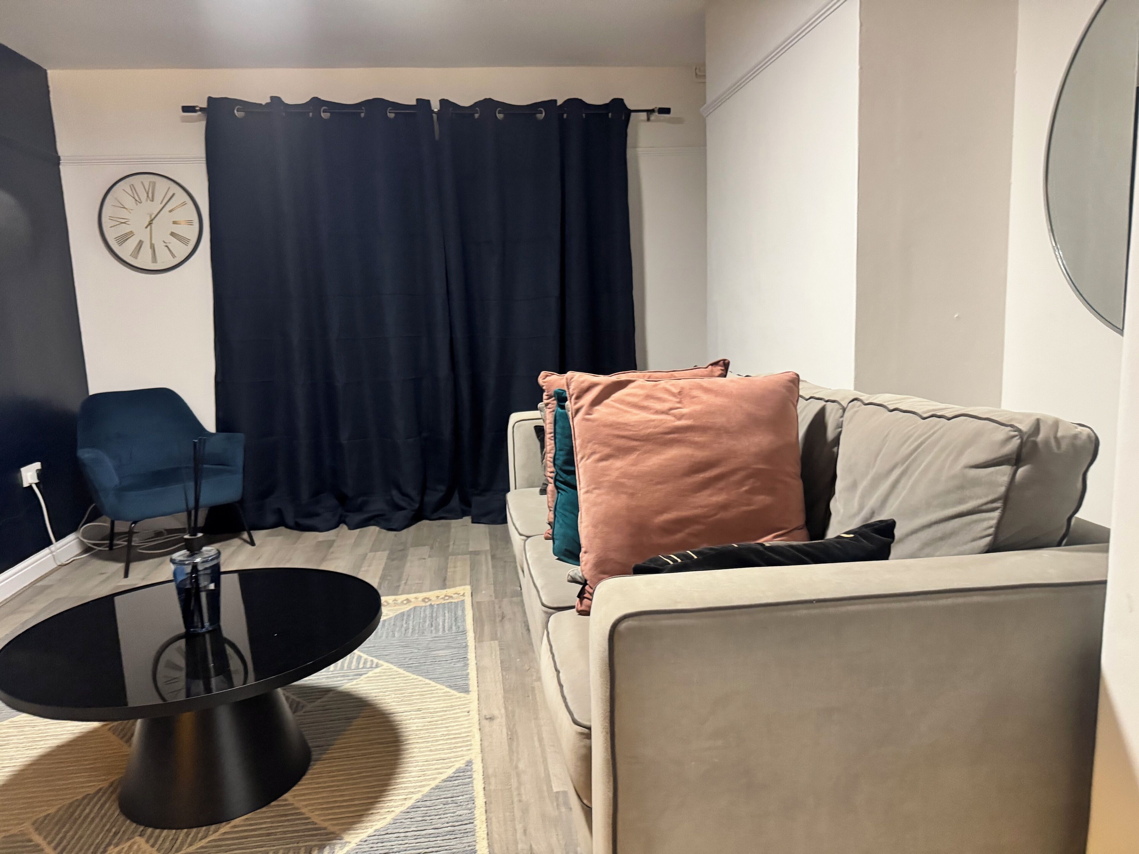 A living room with a sofa, a blue chair, a round coffee table, a clock, and a dark curtain.