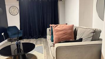 A living room with a sofa, a blue chair, a round coffee table, a clock, and a dark curtain.