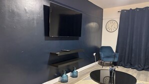 A modern living room with a flat-screen TV mounted on the wall, a round black coffee table, a blue armchair, and a clock on the wall. - Corporate 3bedroom home with parking (Erith)