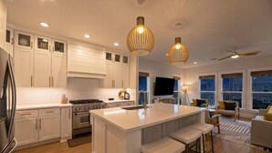 Cocina moderna con armarios blancos, una isla central, taburetes de bar, iluminación colgante y vistas a la ciudad a través de amplios ventanales.
