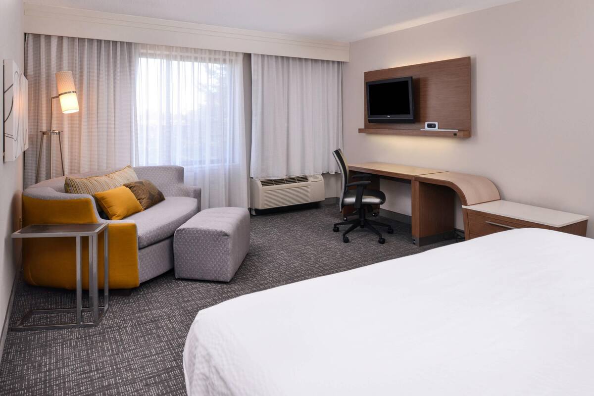 Habitación de hotel con cama, escritorio, silla, sofá, reposapiés, lámpara y televisor montado en la pared.