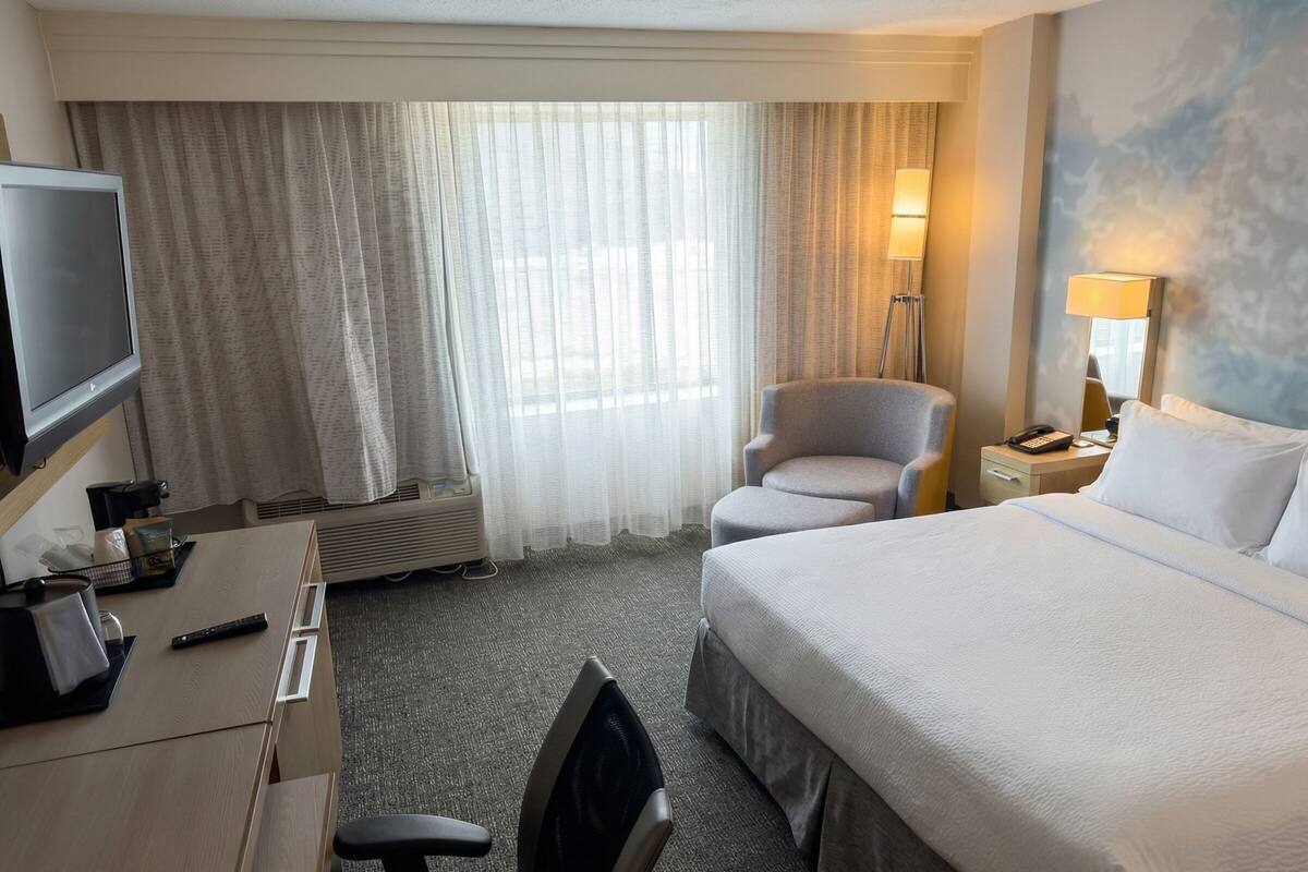 Habitación de hotel con cama, escritorio con televisor, silla, lámpara y ventana con cortinas.