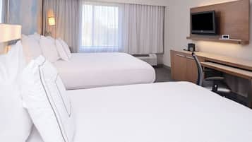 A hotel room with two beds, a desk, a flat-screen TV, and a window with curtains.