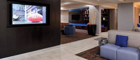 A modern hotel lobby with a flat-screen TV, a seating area with a sofa and ottoman, a patterned carpet, and a bookshelf.
