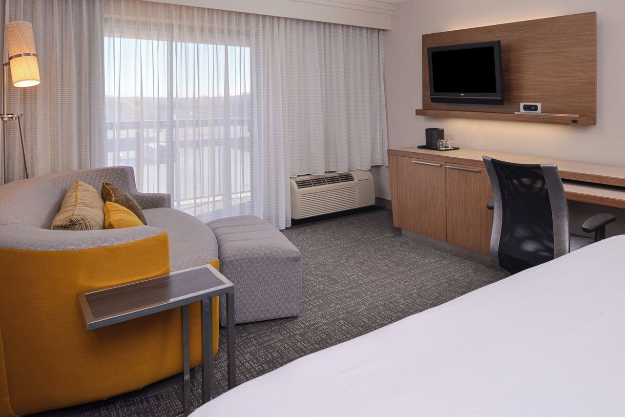 A hotel room with a sofa, ottoman, desk, chair, TV, and air conditioning unit.