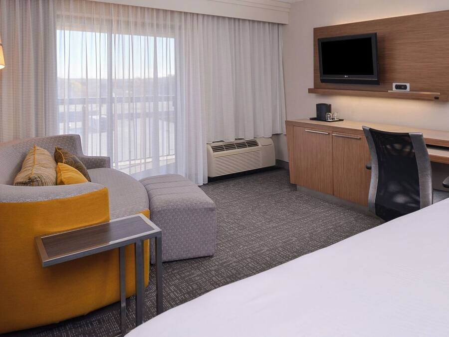 A hotel room with a sofa, ottoman, desk, chair, TV, and air conditioning unit.