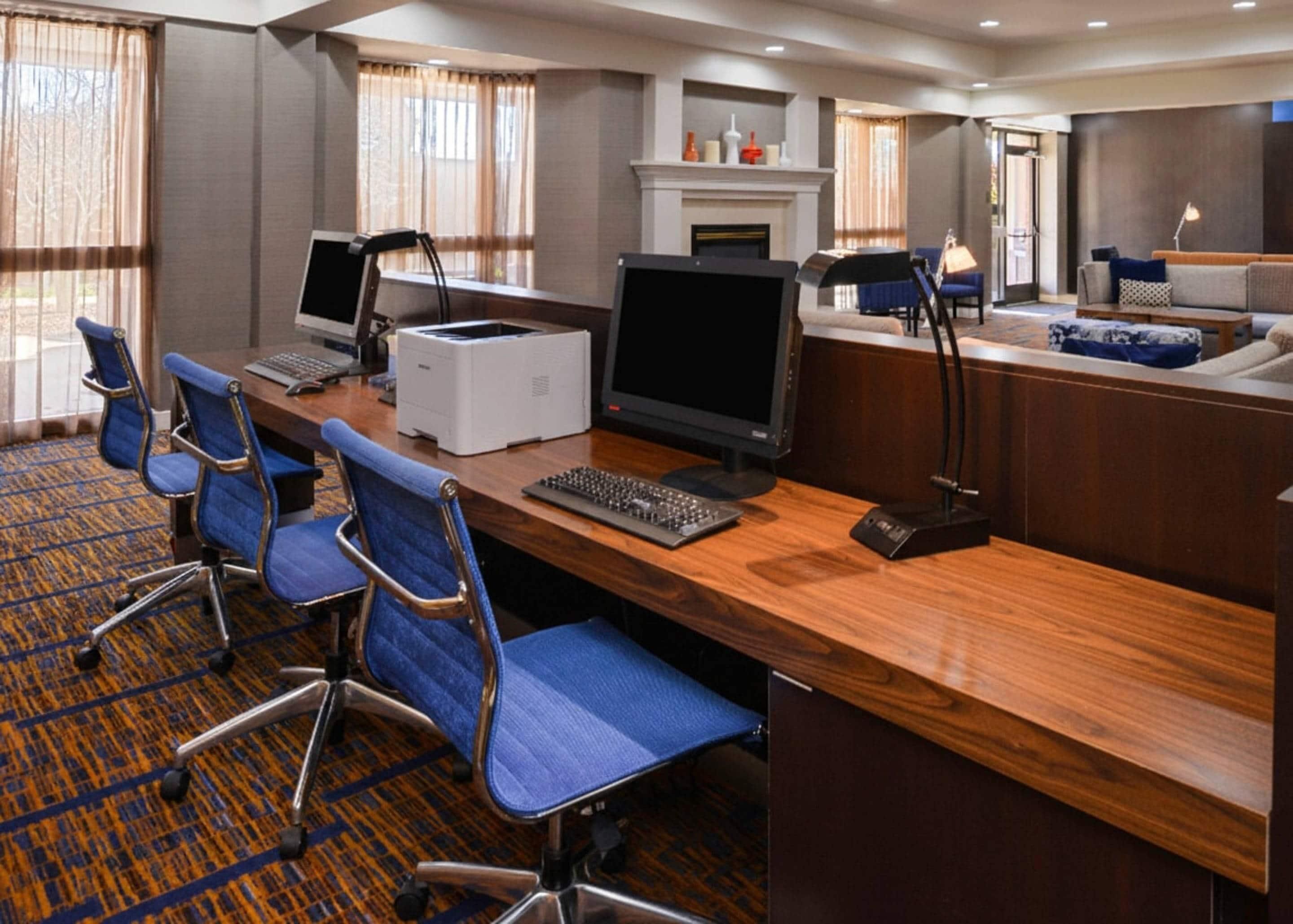 Ein Hotelzimmer mit einem Holztisch, zwei Computern, einem Drucker und einem Kamin. Vier blaue Stühle und ein Sofa sind ebenfalls vorhanden.