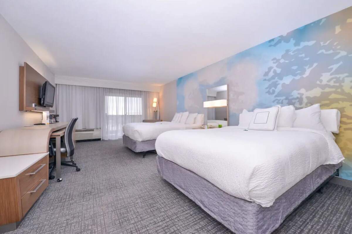 A hotel room with two beds, a desk, a chair, a TV, and a large window with curtains.
