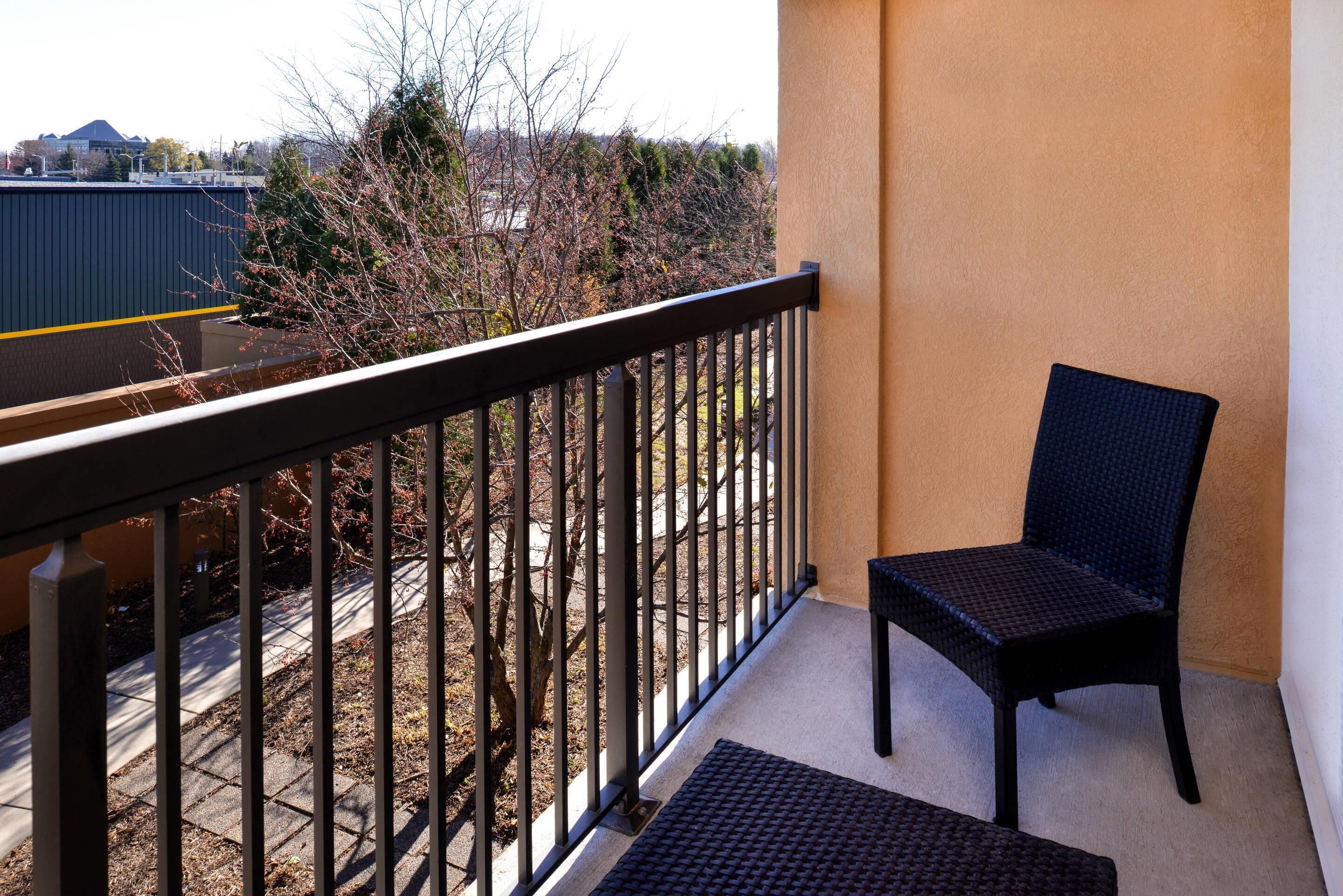 A balcony with a chair, a black metal railing, and a view of trees and a building in the background.