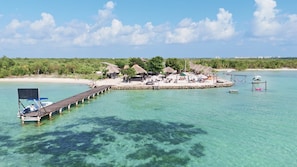 Ett semesterparadis med en brygga som sträcker sig ut i det klara, turkosfärgade vattnet, omgivet av frodig grönska och byggnader med halmtak.