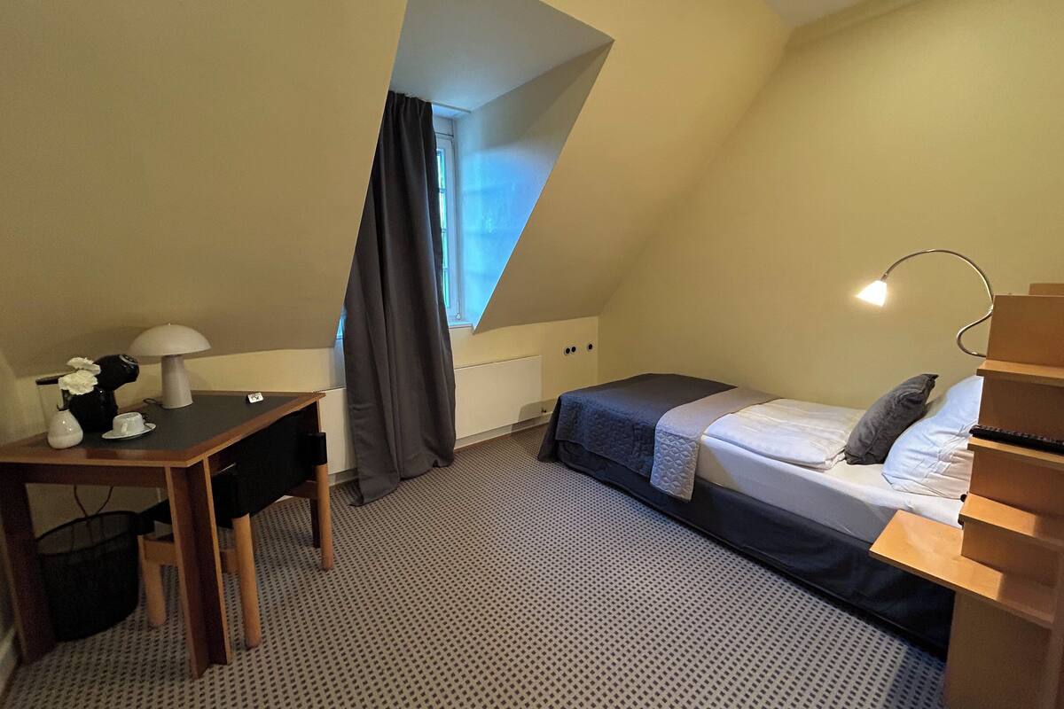 A small, triangular attic bedroom with a bed, desk, lamp, and a window with curtains.