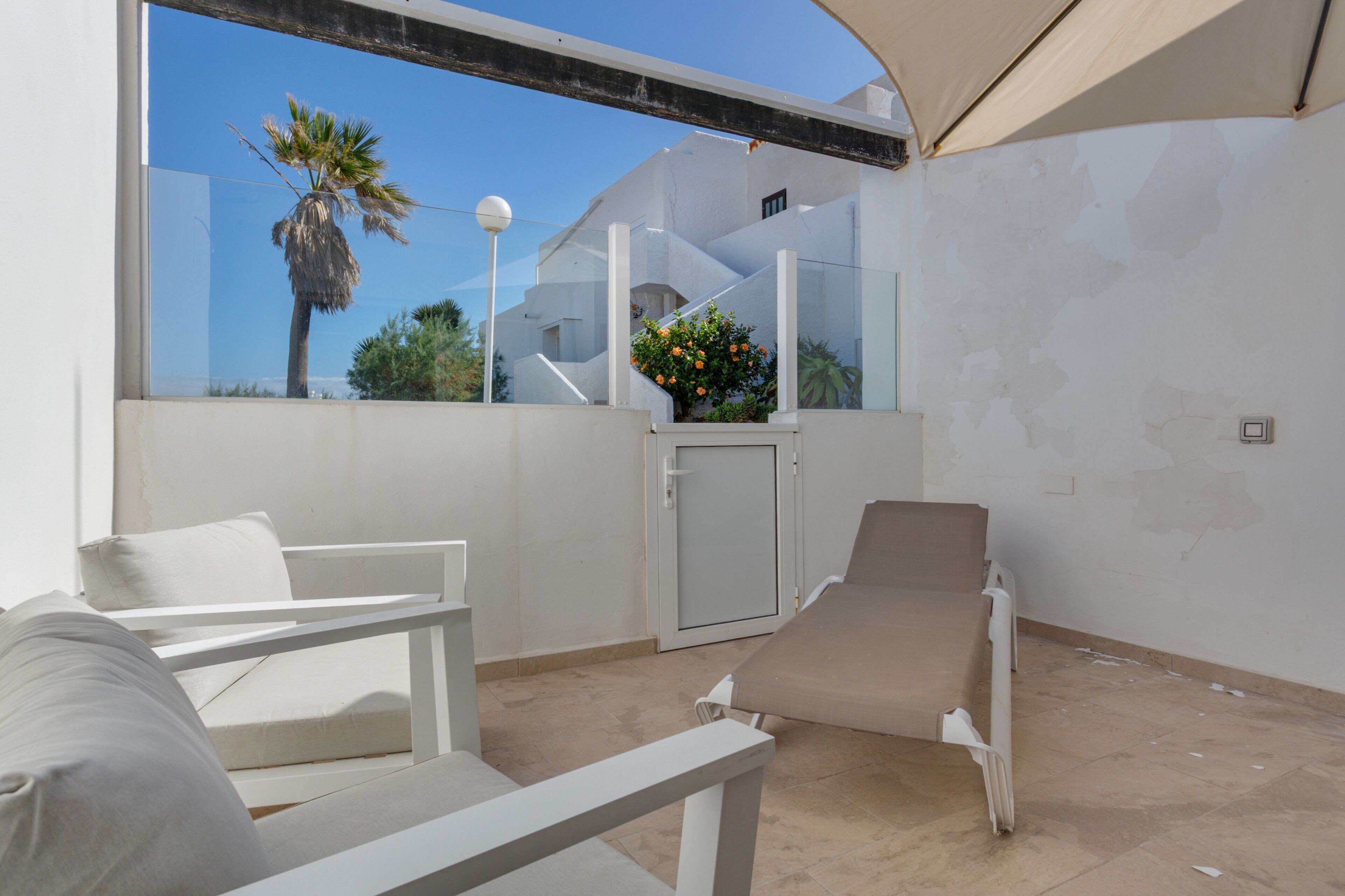 Un espacio al aire libre iluminado por el sol con una silla blanca, una tumbona y una puerta que da a un balcón cubierto.