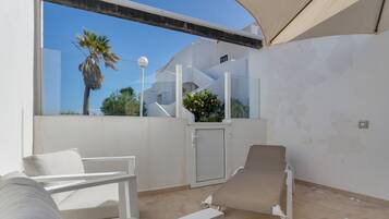 Un espacio al aire libre iluminado por el sol con una silla blanca, una tumbona y una puerta que da a un balcón cubierto.