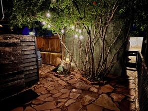A stone pathway leading to a wooden structure with a bench and a tree with hanging lights. - The Dollhouse (Page)