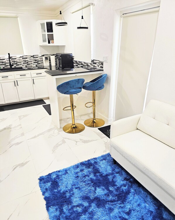 A modern kitchen with white cabinets, a black countertop, and a built-in oven. There are two blue bar stools with gold legs and a white sofa with a blue shag rug.