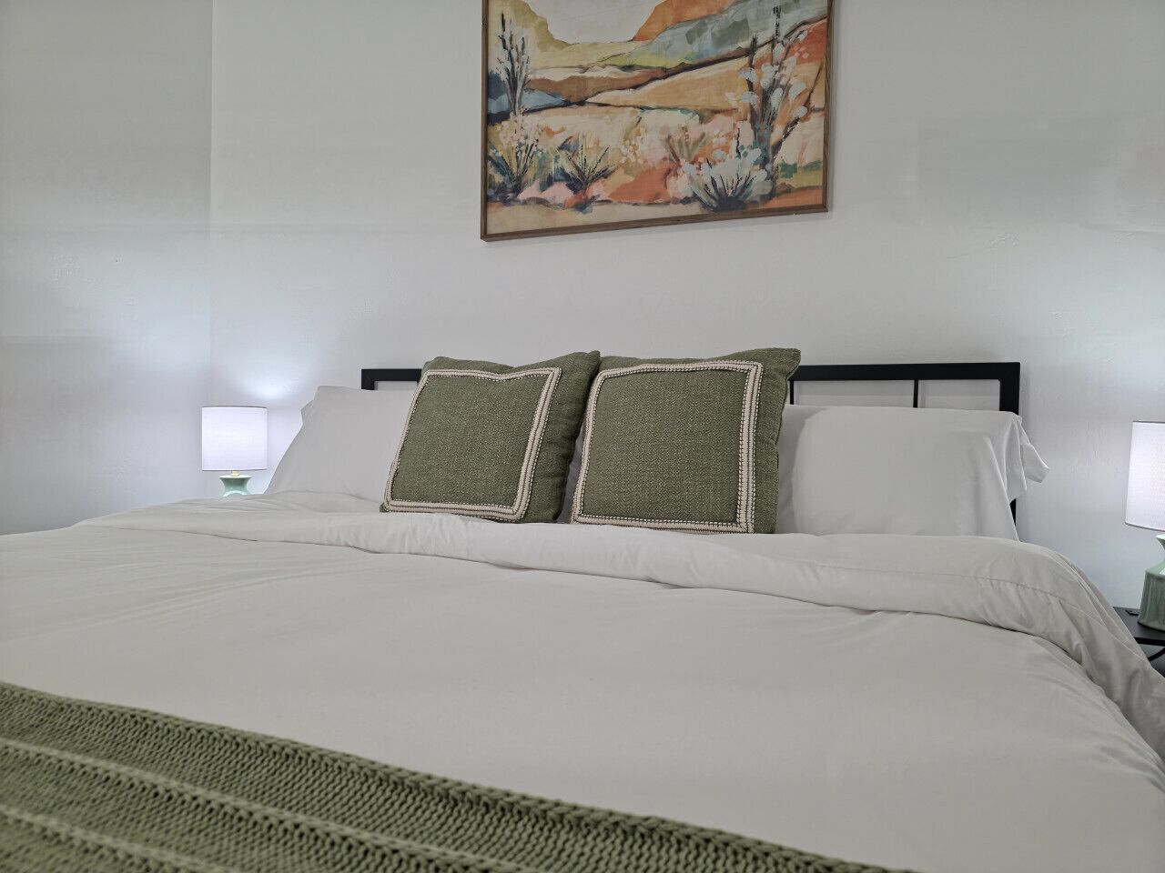 Una cama bien hecha con sábanas blancas y dos almohadas, un cuadro en la pared y dos lámparas de noche.