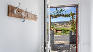 Una puerta que da a un balcón con vistas a un paisaje rural, un perchero de madera en la pared con llaves, y una reja metálica.