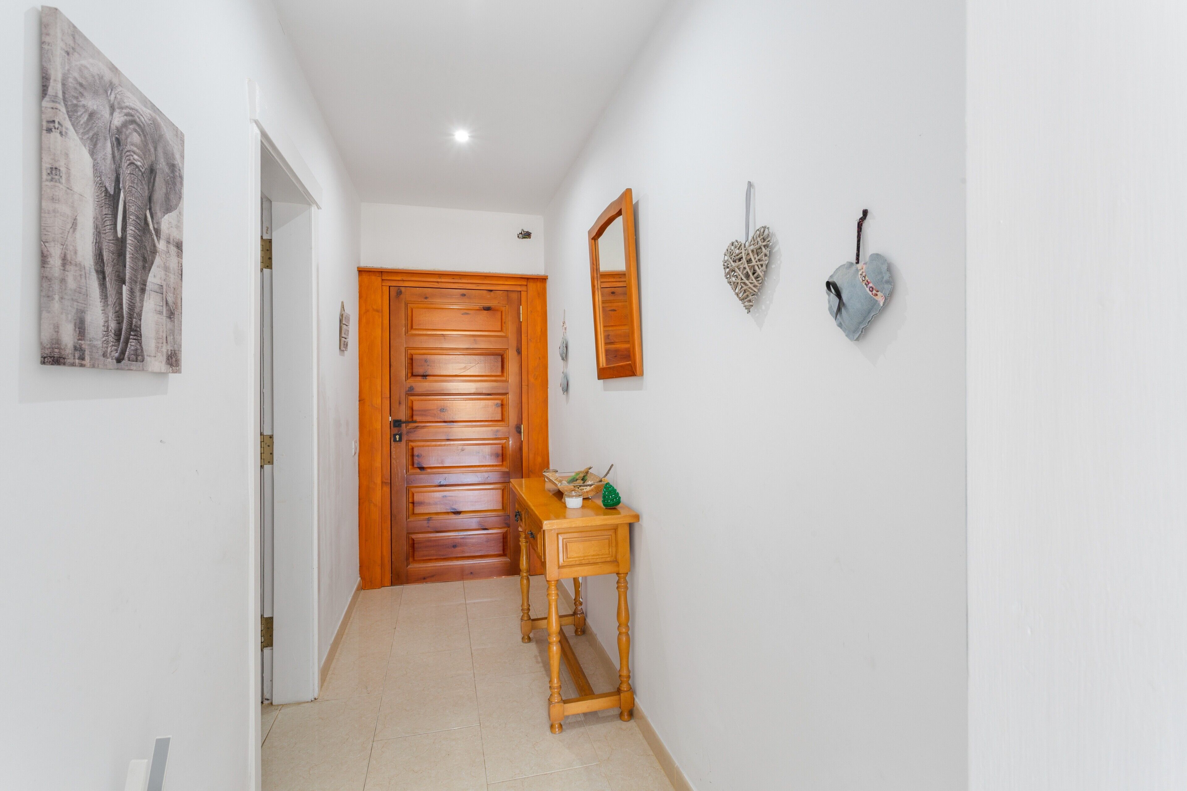Un couloir avec une porte en bois, une petite table en bois sur laquelle se trouve un panier, un miroir et une décoration en forme de cœur accrochée au mur.