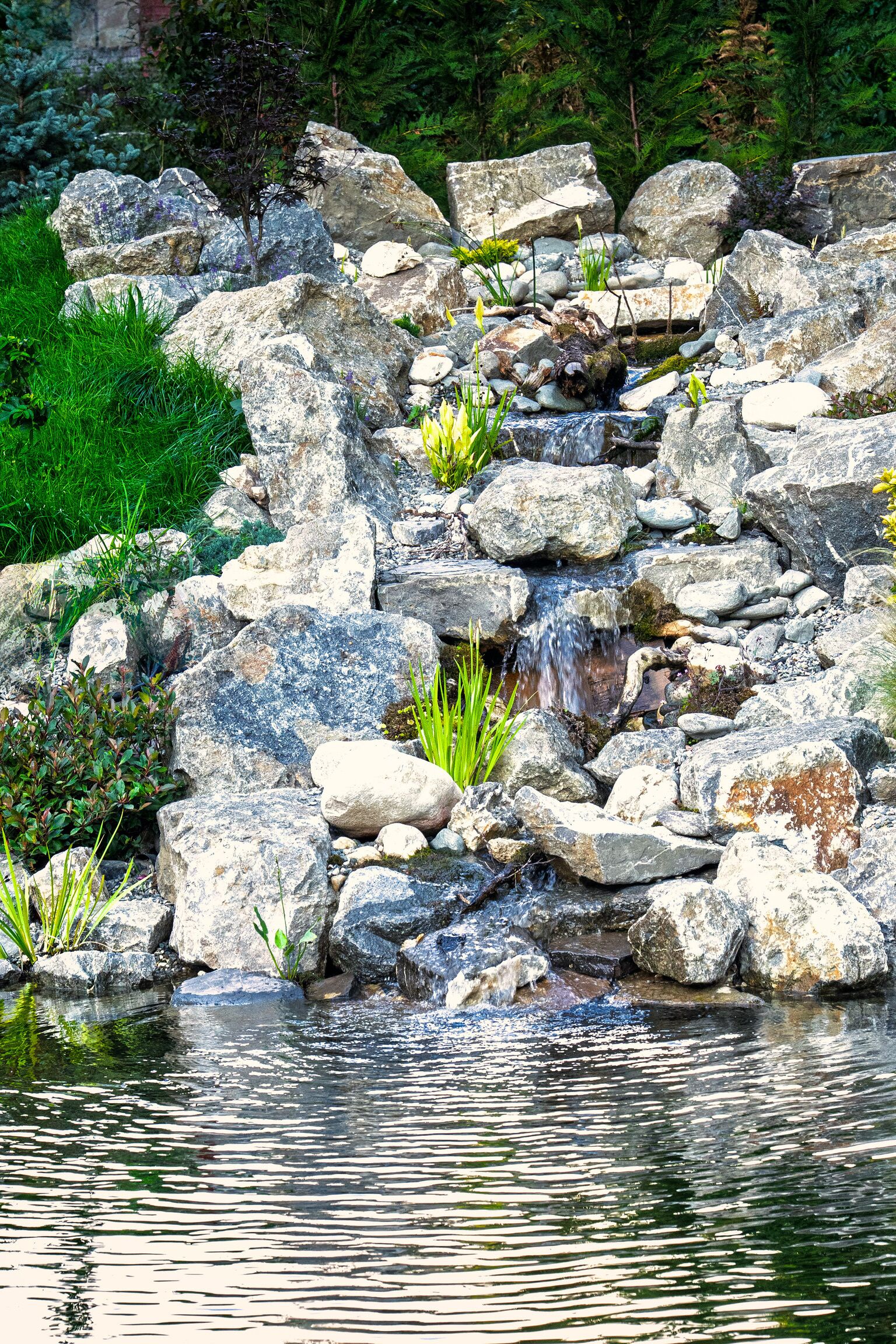 A natural rock formation with a small stream flowing over it, surrounded by greenery.