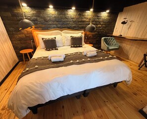 A neatly made bed with white linens and a gray throw blanket, a stone wall, and a wooden floor. - Ocean View Hot Tub Suites (Pichilemu)