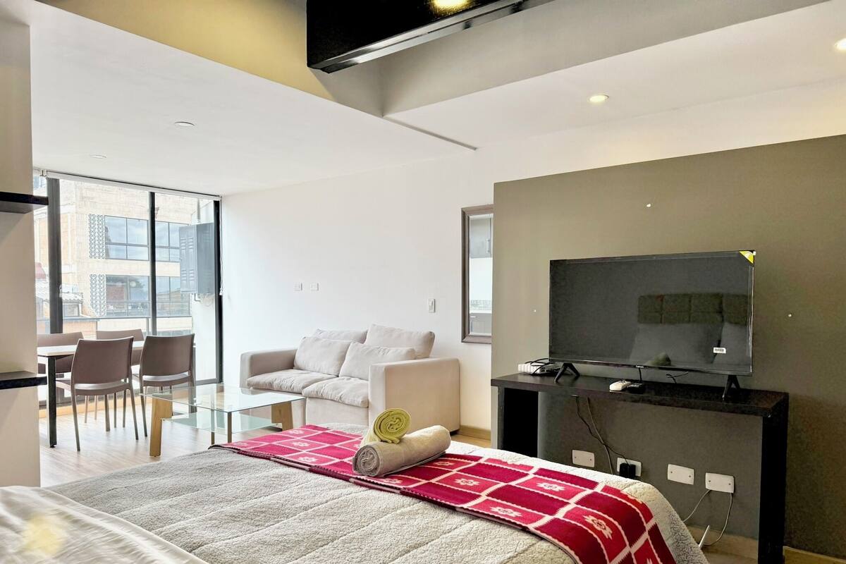A modern hotel room with a large bed, a TV, a desk, and a seating area with a sofa and chairs.