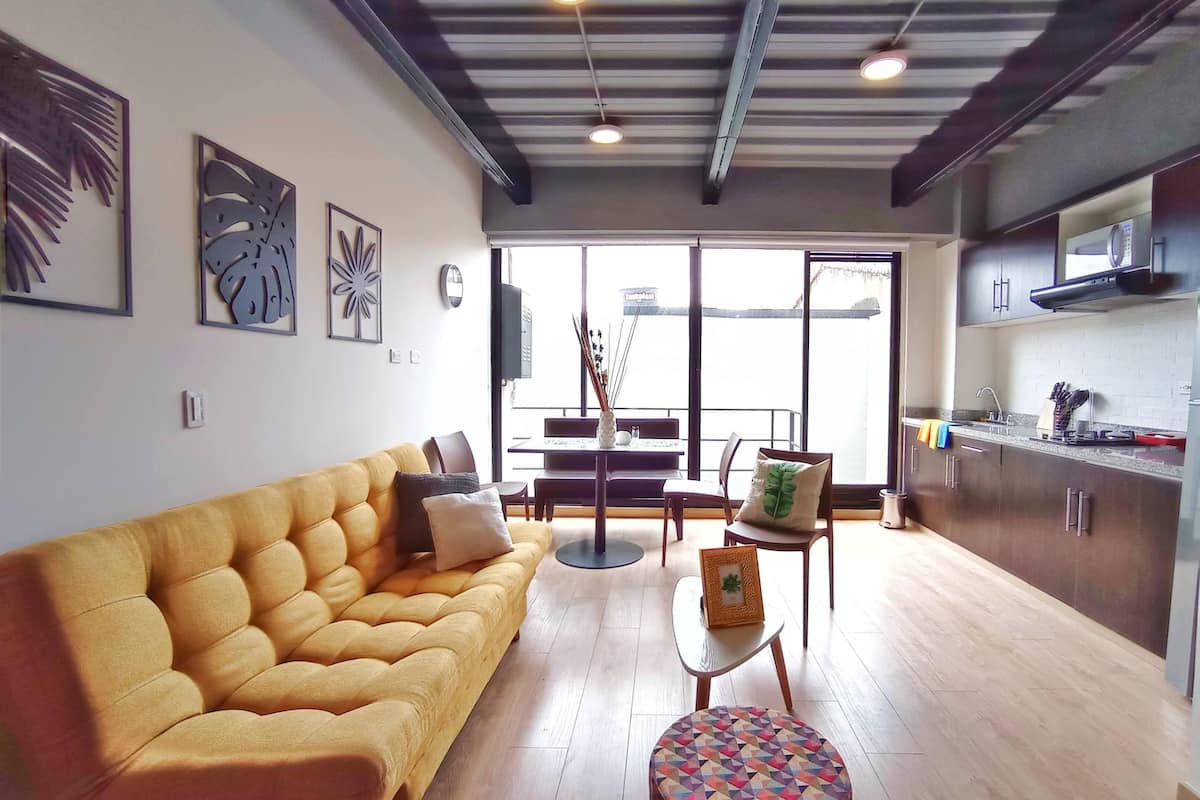 A modern living room with a sofa, a small table, and a colorful ottoman. There is a kitchen area with cabinets and a microwave.