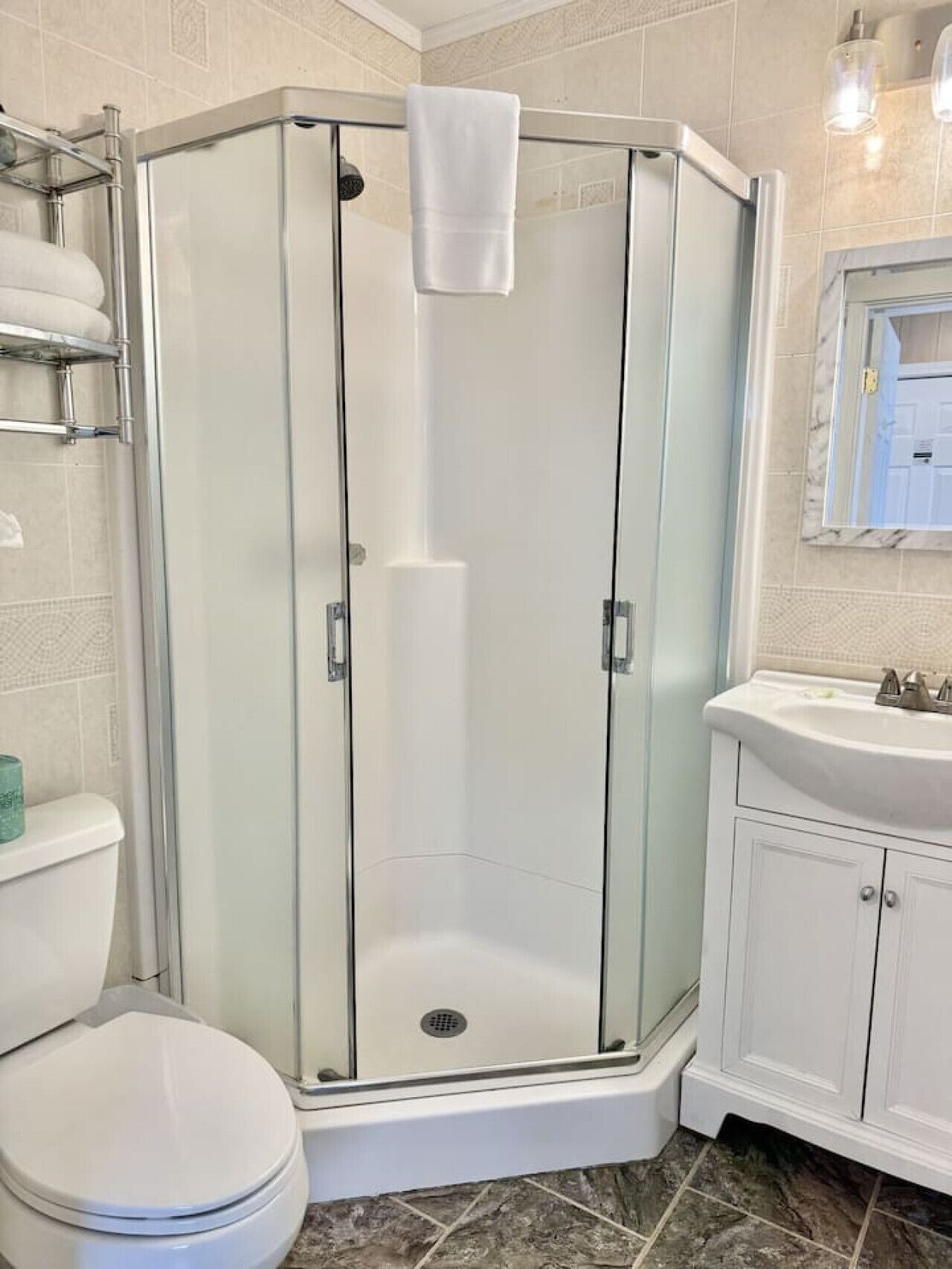 Un espace douche d’angle avec une porte en verre, une serviette blanche accrochée à la porte, des toilettes blanches et un lavabo avec un miroir au-dessus.