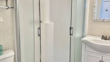 Un espace douche d’angle avec une porte en verre, une serviette blanche accrochée à la porte, des toilettes blanches et un lavabo avec un miroir au-dessus.