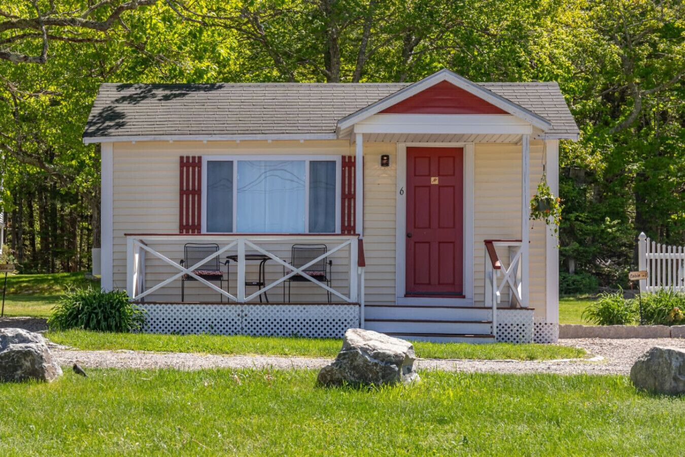 A small, single-story cabin with a red door, white-framed windows, and a covered porch with metal chairs.