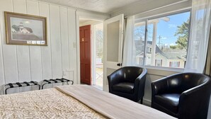 Ett rum med en säng, två svarta stolar, en inramad tavla på väggen och ett fönster med utsikt över hus.