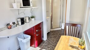 Ett kompakt kök med mikrovågsugn, kaffebryggare och ett litet handfat. Här finns en papperskorg och ett matbord i trä med en krukväxt.