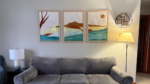 A grey sofa in a room with three framed artworks on the wall, a floor lamp, and a side table with a lamp.