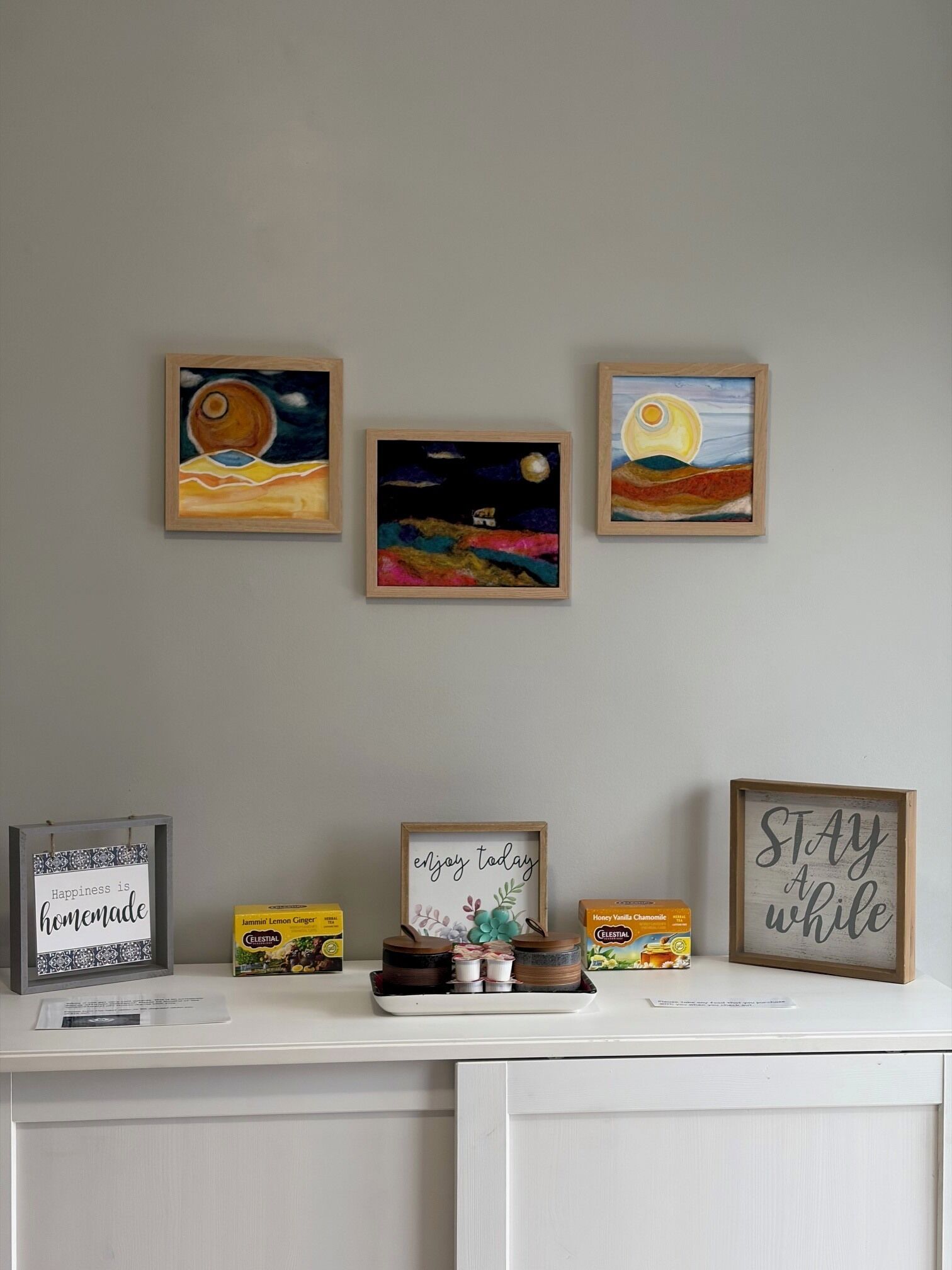 Een muur met drie ingelijste schilderijen die nachttaferelen voorstellen, een plank met potten en een bordje met de tekst 'Geniet van vandaag', en een bordje waarop staat 'Geluk is zelfgemaakt'.