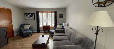 Een woonkamer met een bank, een bijzettafeltje en een lamp. Er is een raam met gordijnen en een deur.