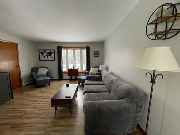 A living room with a sofa, a coffee table, and a lamp. There is a window with curtains and a door.
