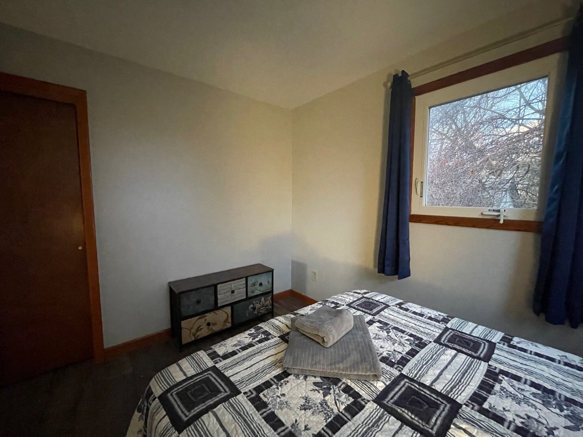 A bedroom with a bed, a dresser, a window with curtains, and a door.