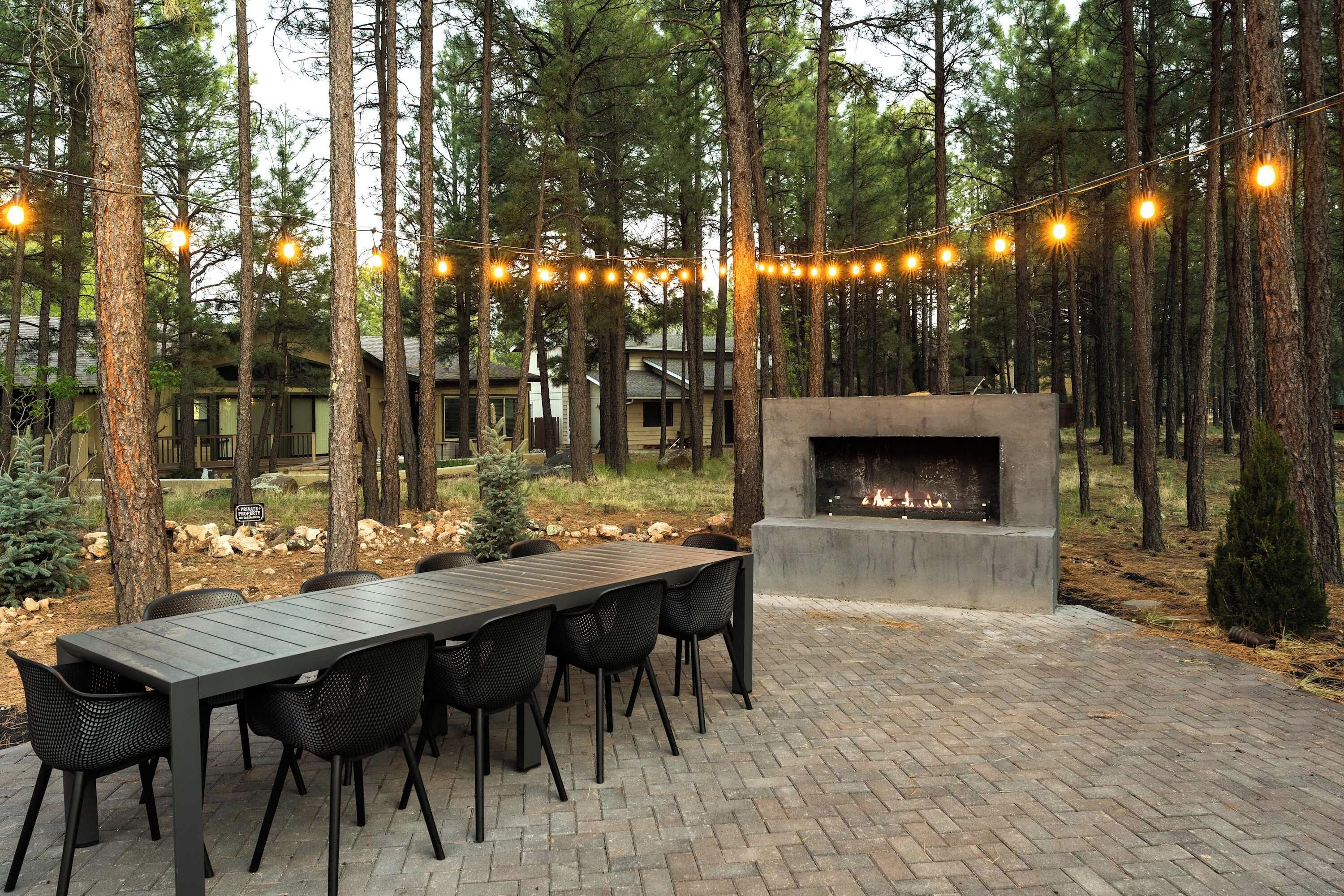 An outdoor patio with a brick floor, a long table, and chairs, surrounded by tall trees and string lights.
