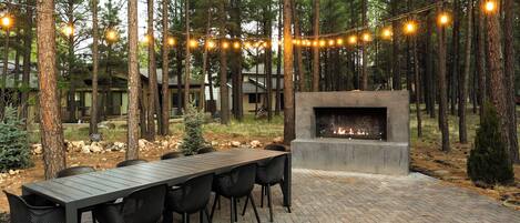 An outdoor patio with a brick floor, a long table, and chairs, surrounded by tall trees and string lights.