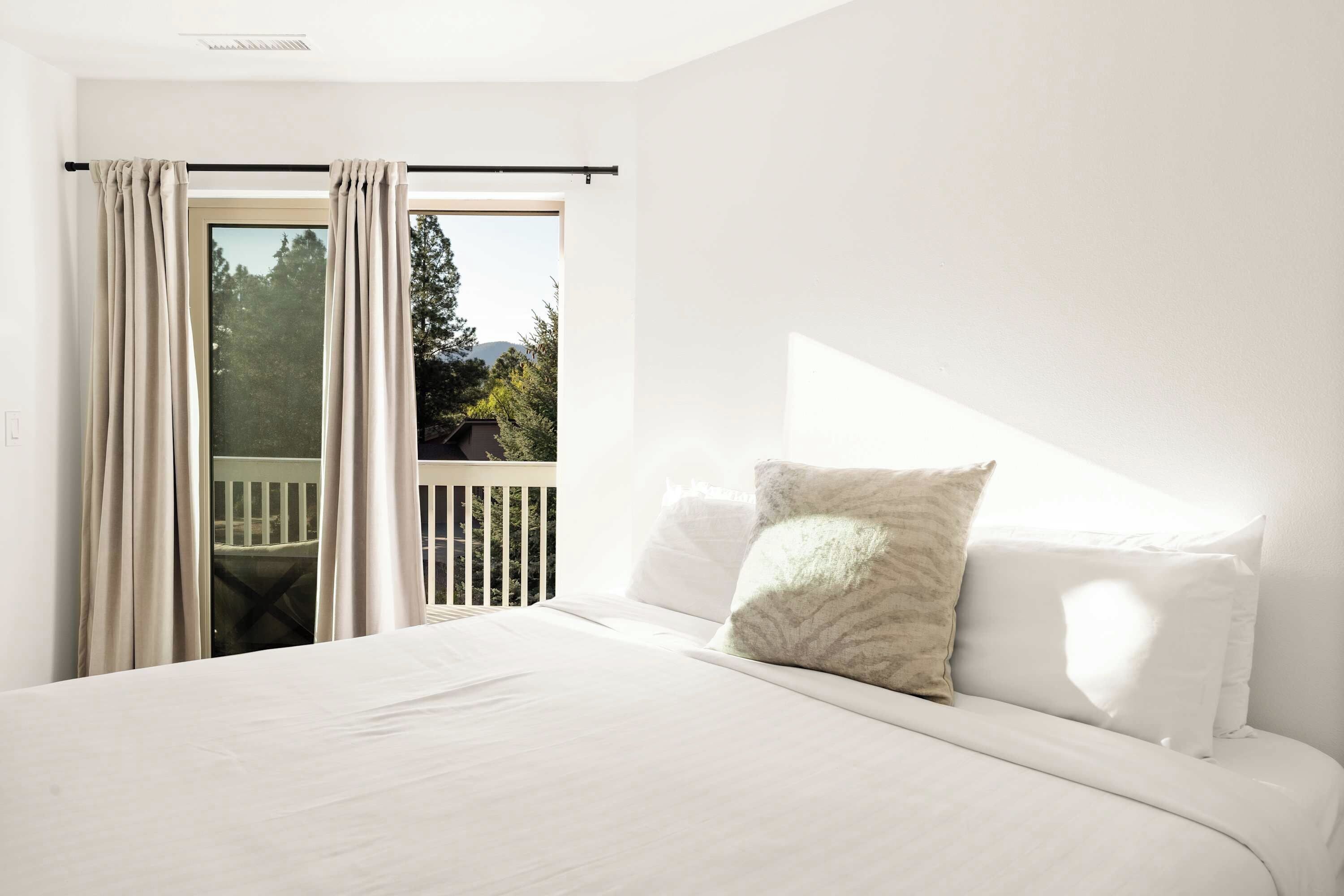 A bedroom with a bed, a window with curtains, and a view of trees and a deck.