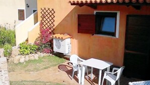Un pequeño patio al aire libre con una mesa y sillas blancas, techo de tejas rojas y una ventana con contraventanas.