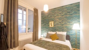 Hotellihuone, jossa on sänky, yöpöytä, lamppu, ikkuna verhoilla ja aaltoilevakuviollinen seinä.