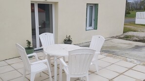 A patio with a round table and four white chairs, potted plants, a sliding glass door, and a small window with a green shutter. - In the quiet countryside, single-storey house (Mouilleron-le-Captif)