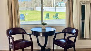 A room with a round table and two chairs, a window with curtains, and a view of a grassy area and a fence. - Gallagher`s Travels Room 12-A (King Bed) (Bar Harbor)