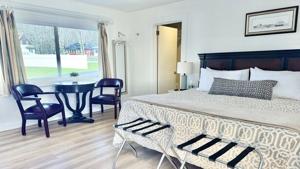 A hotel room with a bed, a desk, two chairs, a small table, and a view of the outside. - Gallagher`s Travels Room 12-A (King Bed) (Bar Harbor)