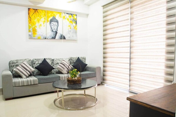 A living room with a grey sofa, black and white pillows, a round coffee table with a basket, and a large window with blinds. - H2o Residences Colombo (Colombo)