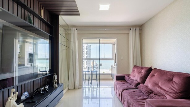 A modern living room with a large flat-screen TV, a plush pink sofa, and a glass door leading to a balcony with a city view. - Apartment Sea View 500 m From the Beach Roo0401 (Itapema)