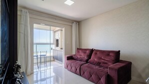 A modern living room with a plush red sofa, a glass door leading to a balcony with a view, and a minimalist decor style. - Apartment Sea View 500 m From the Beach Roo0401 (Itapema)
